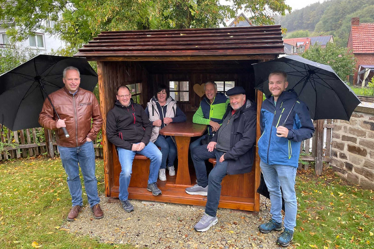 Sechs Personen posieren bei Regen an und in einer hölzernen Schutzhütte; zwei Männer links und rechts halten geöffnete, schwa