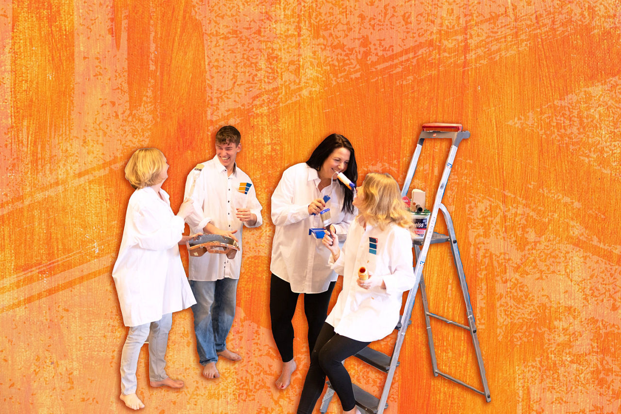 Drei Frauen und ein Mann stehen mit Pinsel und Farbe vor einer orangenen Wand.