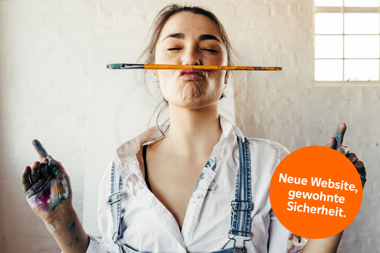 Eine junge Frau steht mit Farbflecken auf der Kleidung und einem Pinsel zwischen Mund und Nase geklemmt in einem Atelier