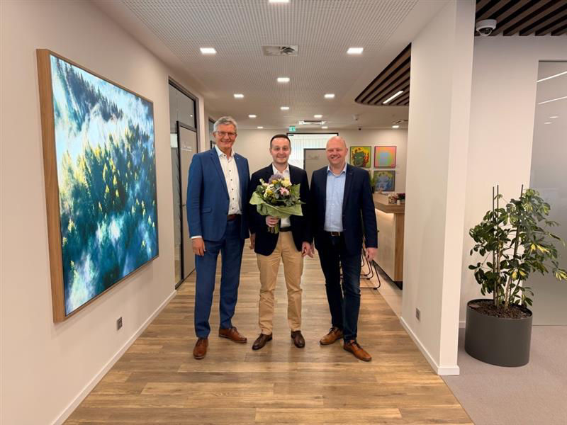 Zwei Männer und ein Mann haben sich zum Foto aufgestellt. Der Mann in der Mitte hat einen Blumenstrauß in der Hand.