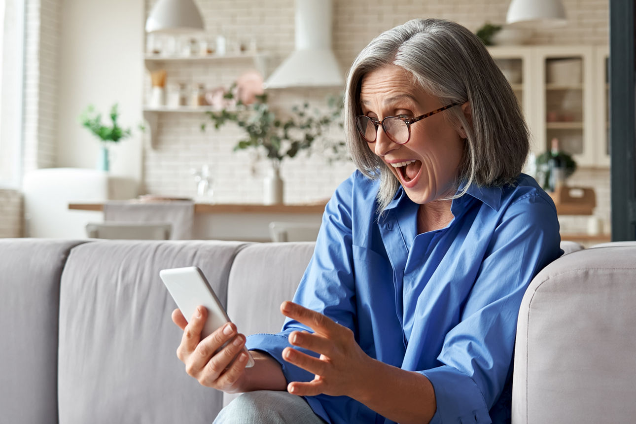 Eine sympathische Dame sitzt auf einem Sofa und schaut lachend auf ihr Smartphone.