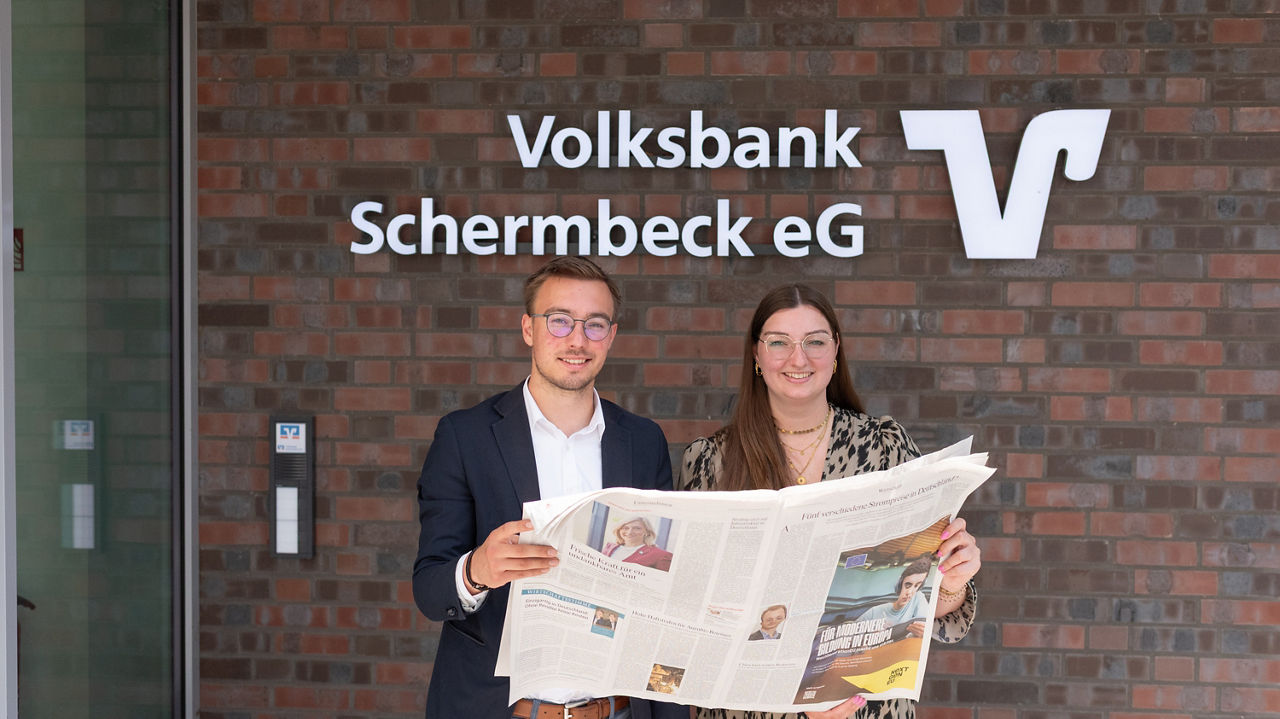 Junger Mann und junge Frau stehen mit Zeitung in der Hand vor dem Bankgebäude der Volksbank Schermbeck