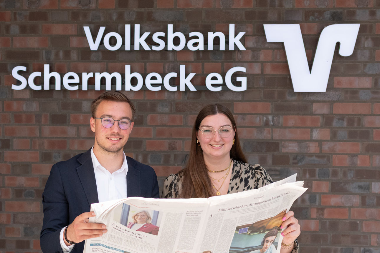 Junger Mann und junge Frau stehen mit Zeitung in der Hand vor dem Bankgebäude der Volksbank Schermbeck