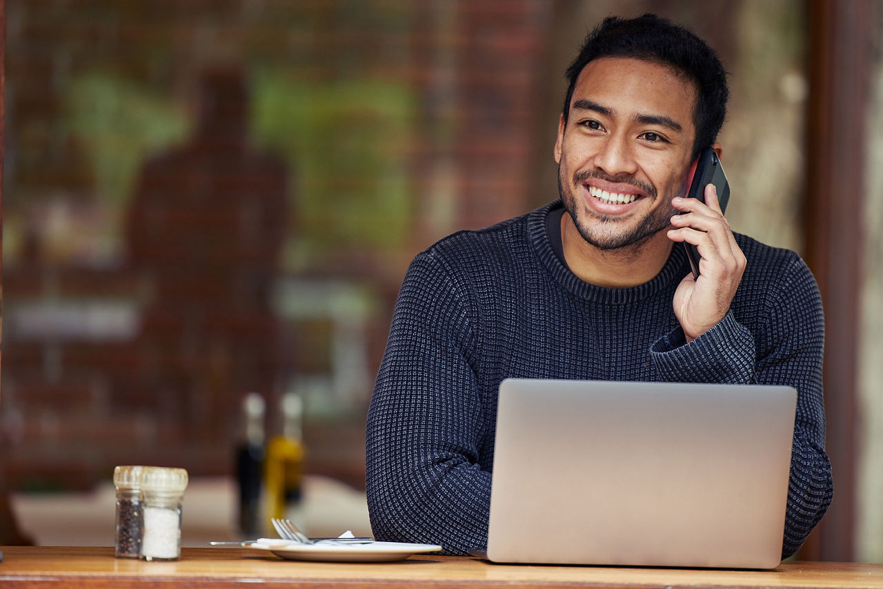 Ein Mann, der in einem Café sitzt und mit seinem Handy telefoniert und seinen Laptop benutzt.