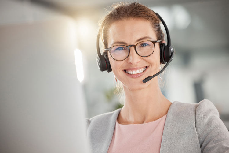 Frau mit Headset schaut lächelnd in die Kamera und sitzt in Businesskleidung an einem Monitor