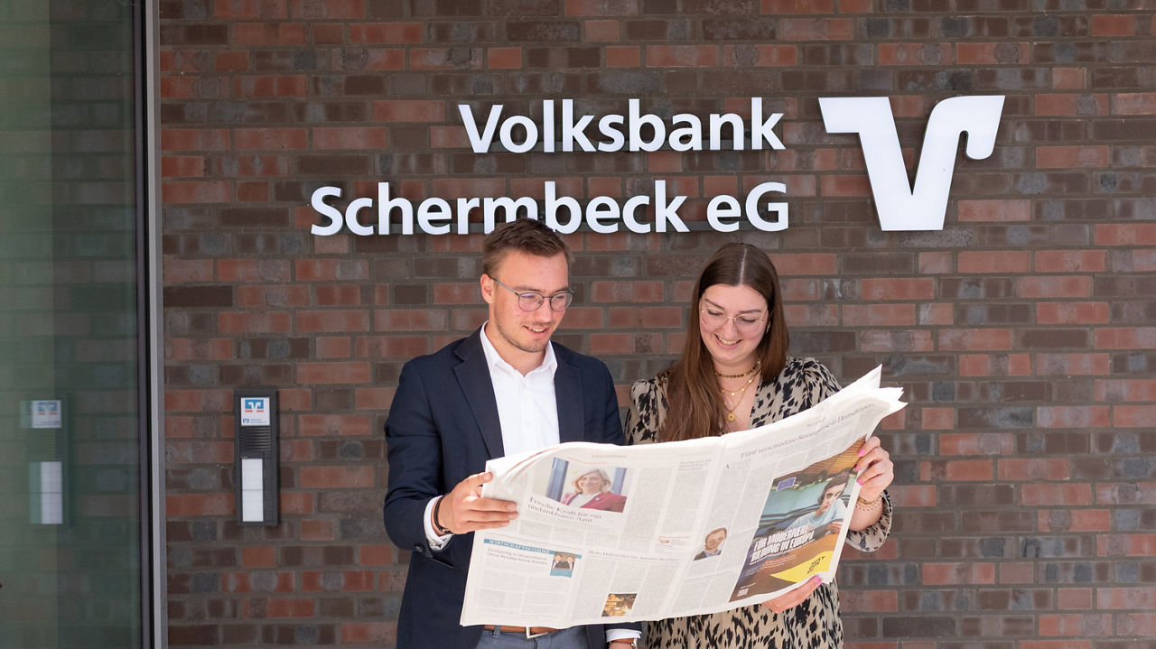 Junger Mann und junge Frau stehen mit Zeitung in der Hand vor dem Bankgebäude der Volksbank Schermbeck