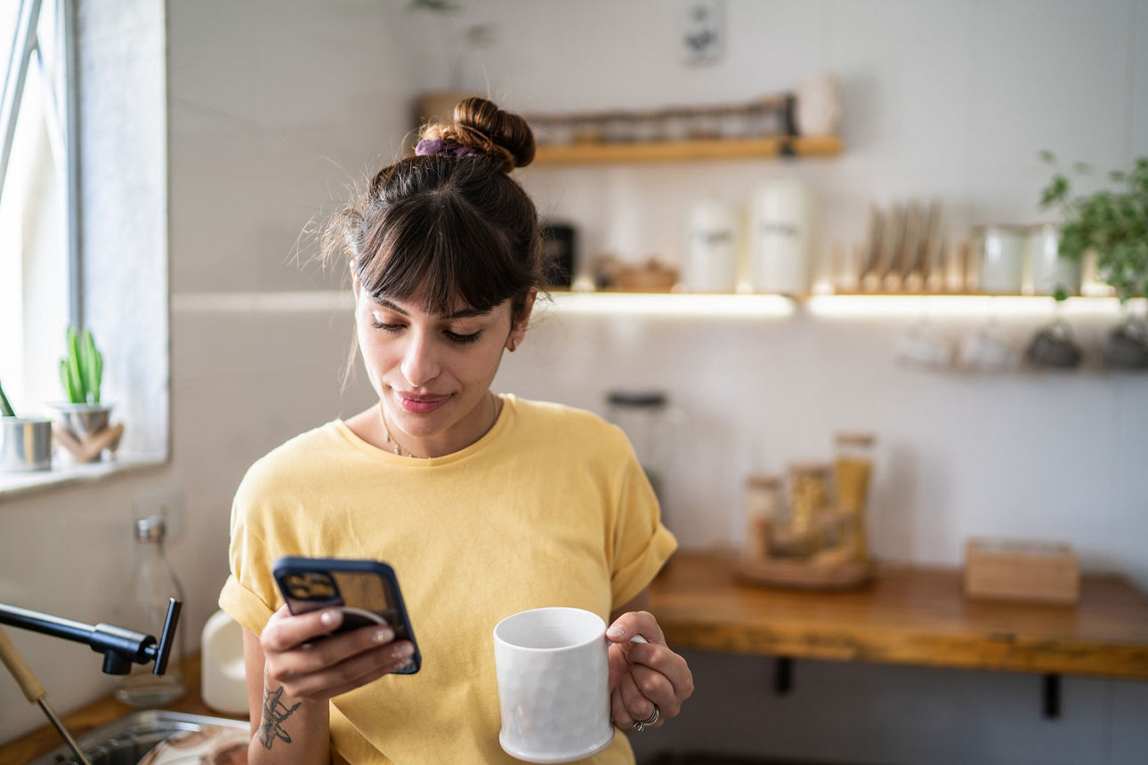 Junge Frau, die das Handy benutzt, während sie zu Hause Kaffee oder Tee trinkt