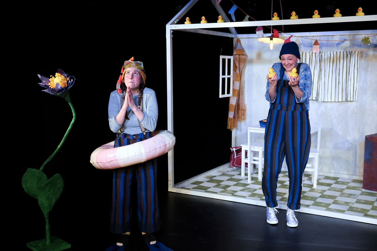 Frau mit Schwimmreif und Frau mit Latzhose stehen vor einem Haus_Nimmerland_Theaterproduktion