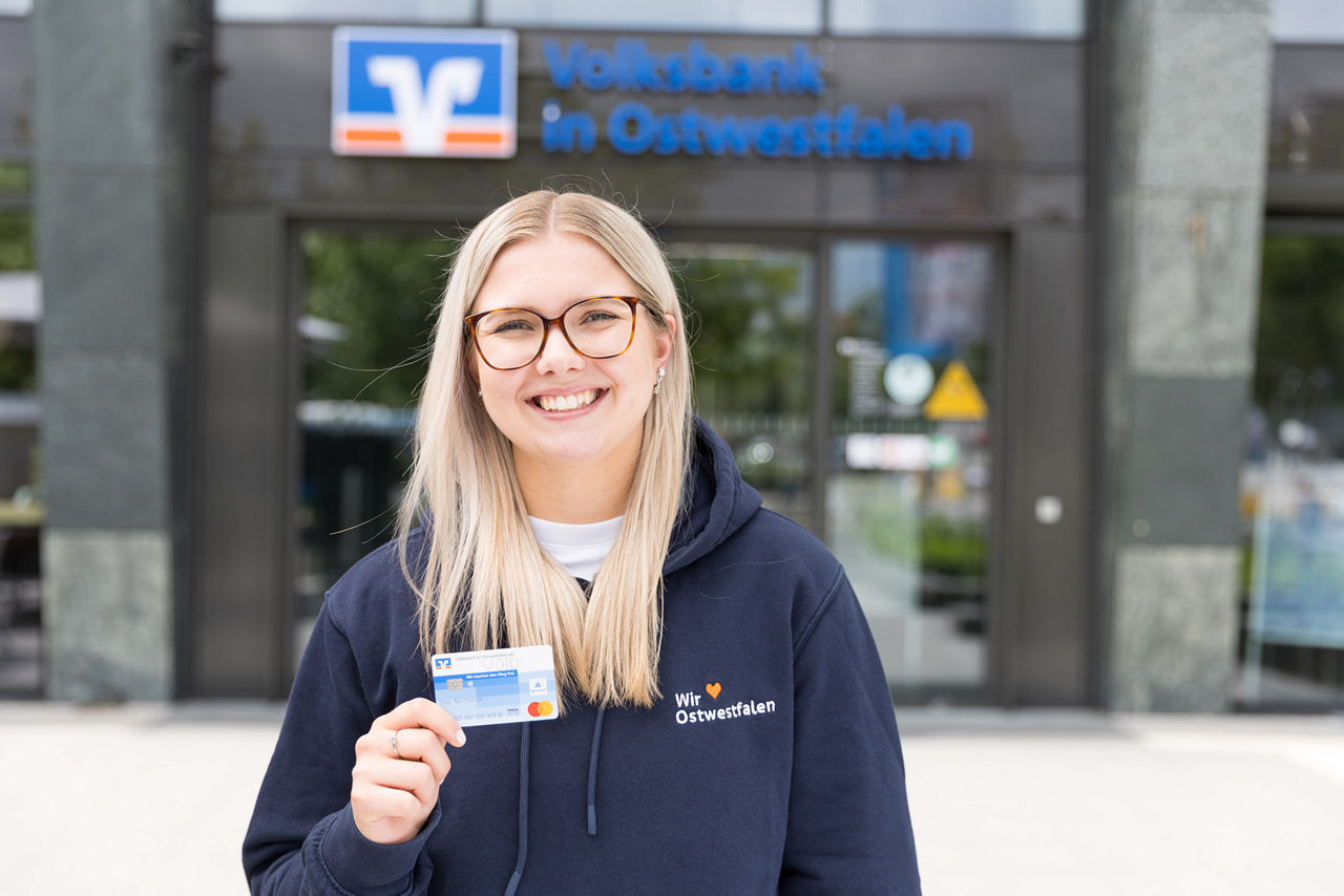Eine junge Frau steht vor einer Geschäftsstelle der Volksbank in Ostwestfalen und hält eine Debitkarte in der Hand.