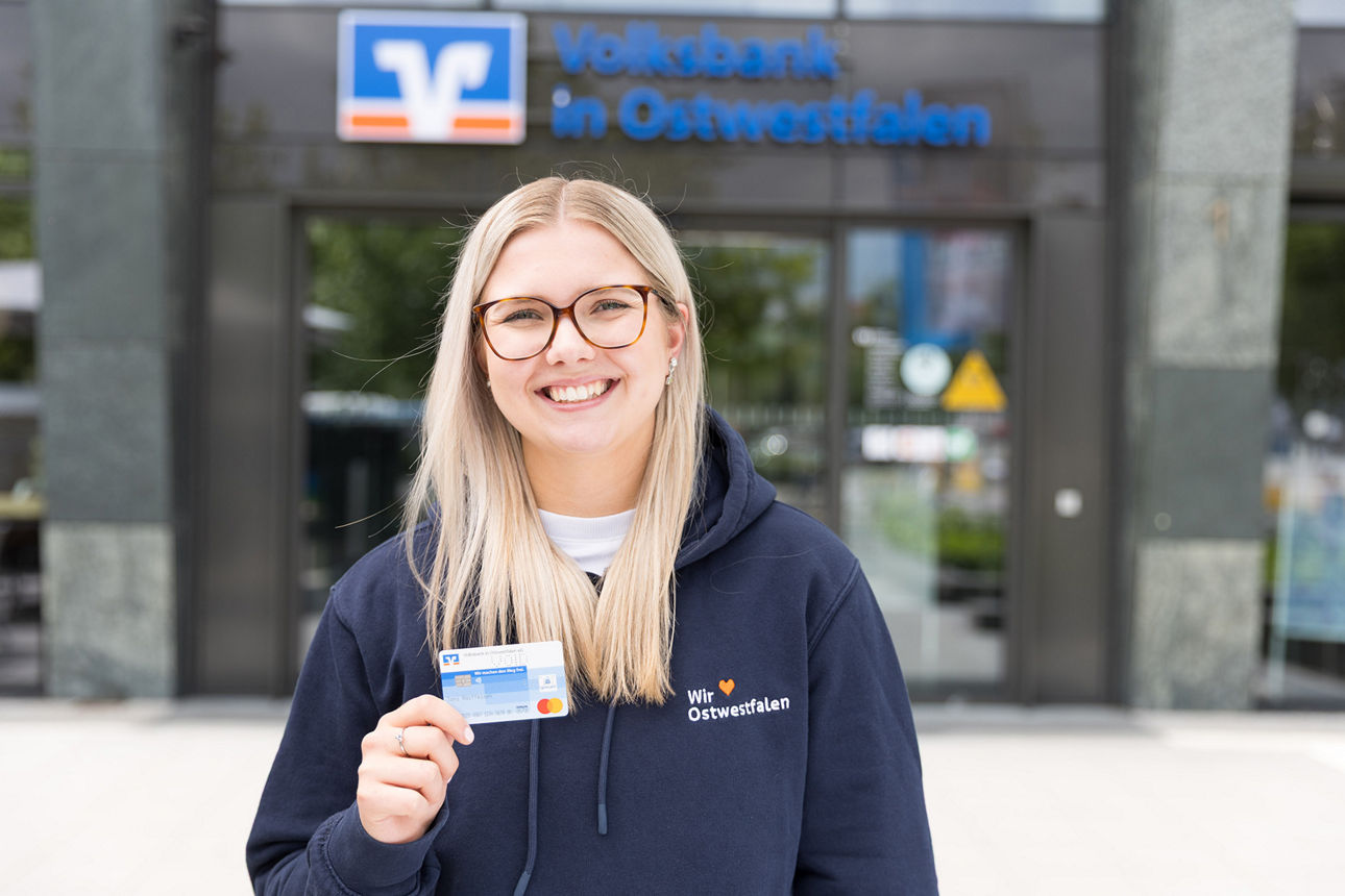 Eine junge Frau steht vor einer Geschäftsstelle der Volksbank in Ostwestfalen und hält eine Debitkarte in der Hand.