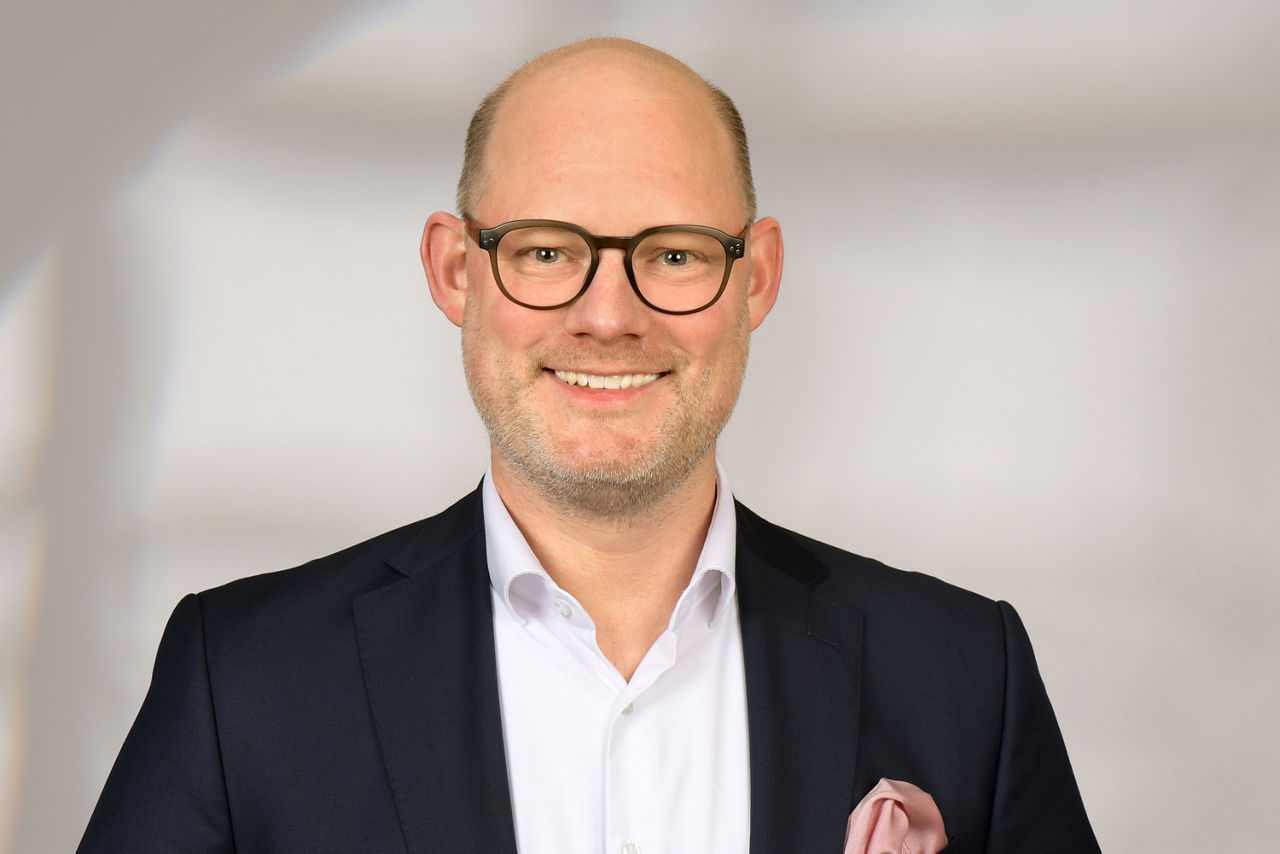 Ein Mann trägt einen dunklen Anzug mit weißem Hemd und einem hellen Einstecktuch vor einem hellen, unscharfen Hintergrund.