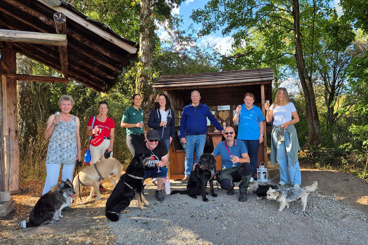 Eine Gruppe von Wanderern posiert mit mehreren Hunden vor einer hölzernen Schutzhütte im Wald und stößt mit Gläsern an.