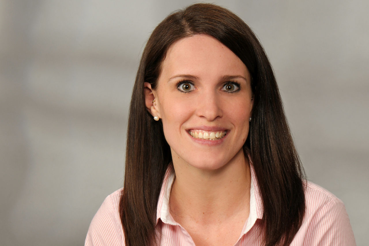 Frau mit langen braunen Haaren, trägt rosa-weiß gestreiftes Hemd, vor neutralem Hintergrund