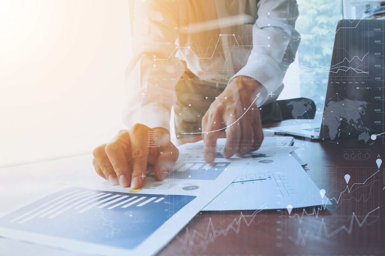 „Zwei Hände, die Papiere auf einem Schreibtisch halten. Im Hintergrund sieht man einen Laptop.