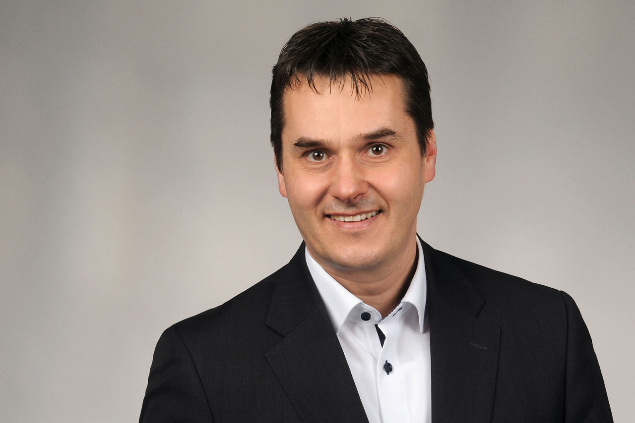 Mann mit dunklem Haar trägt schwarzen Anzug und weißes Hemd mit schwarzer Knopfleiste vor neutralem Hintergrund