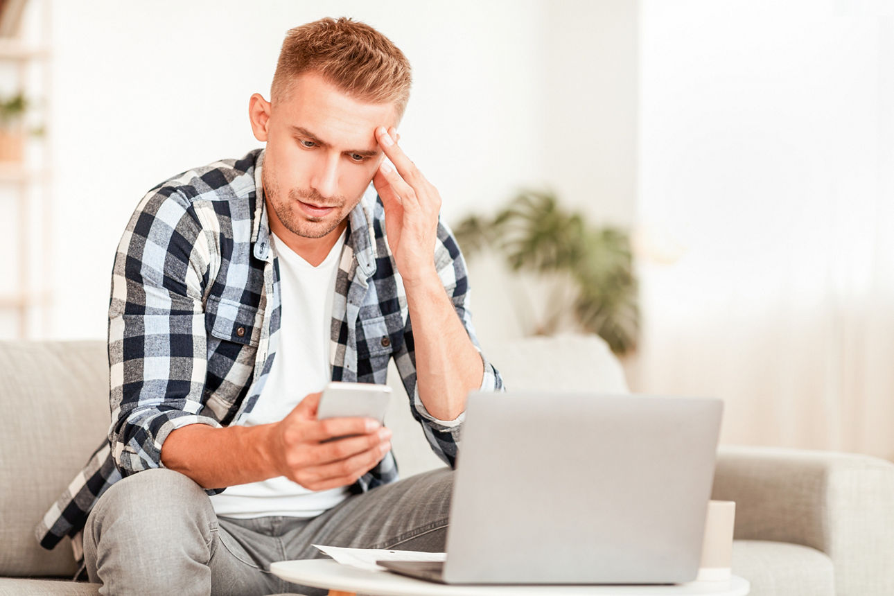 Besorgter Mann sitzt grübelnd auf der Couch vor dem Laptop mit Handy in der Hand.
