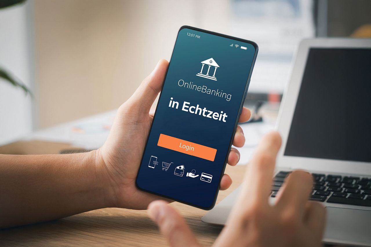 Eine Hand hält ein Smartphone. Auf dem Display steht "Online- Banking in Echtzeit. Im Hintergrund ist ein Laptop angedeutet und eine weitere Hand im Vordergrund, die auf das Smartphone ausgerichtet ist.