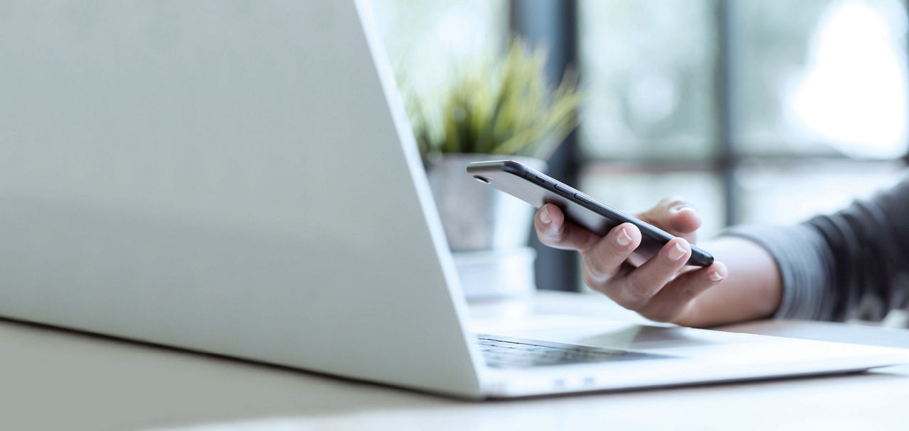 Es steht ein Laptop auf dem Tisch und eine Hand hält ein Smartphone