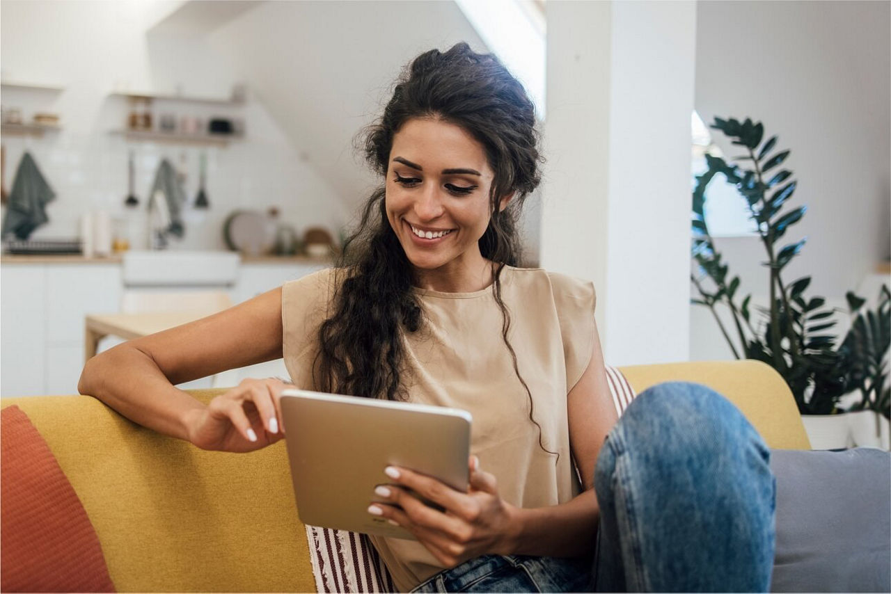 Eine Frau sitzt lächelnd auf dem Sofa mit einem Tablet in der Hand