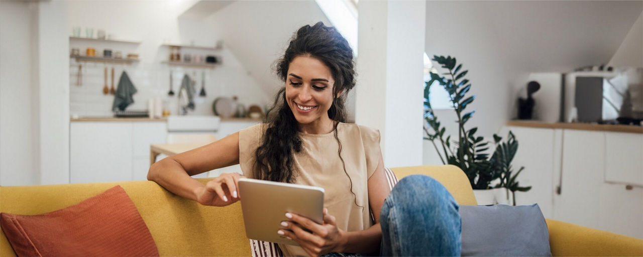 Eine Frau sitzt lächelnd auf dem Sofa mit einem Tablet in der Hand