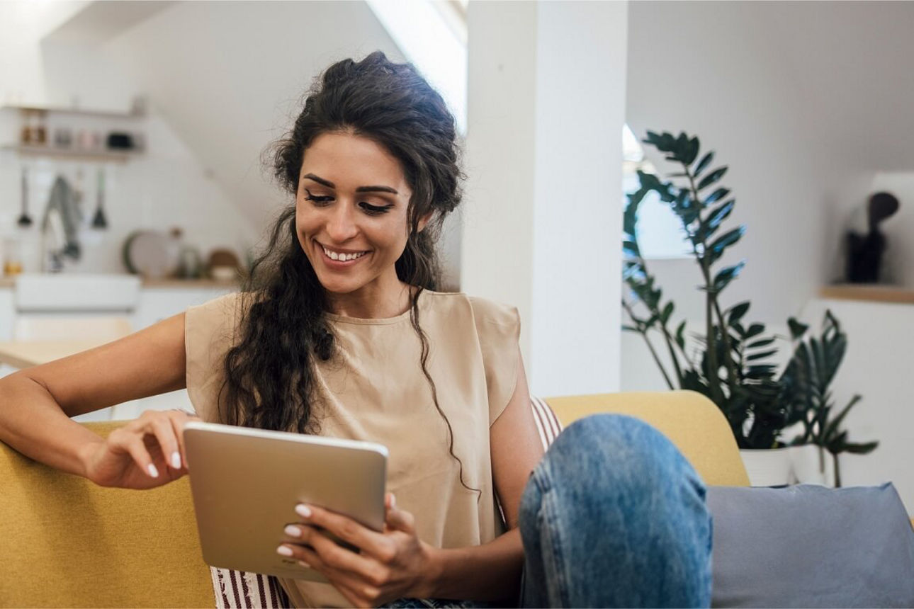Eine Frau sitzt lächelnd auf dem Sofa mit einem Tablet in der Hand