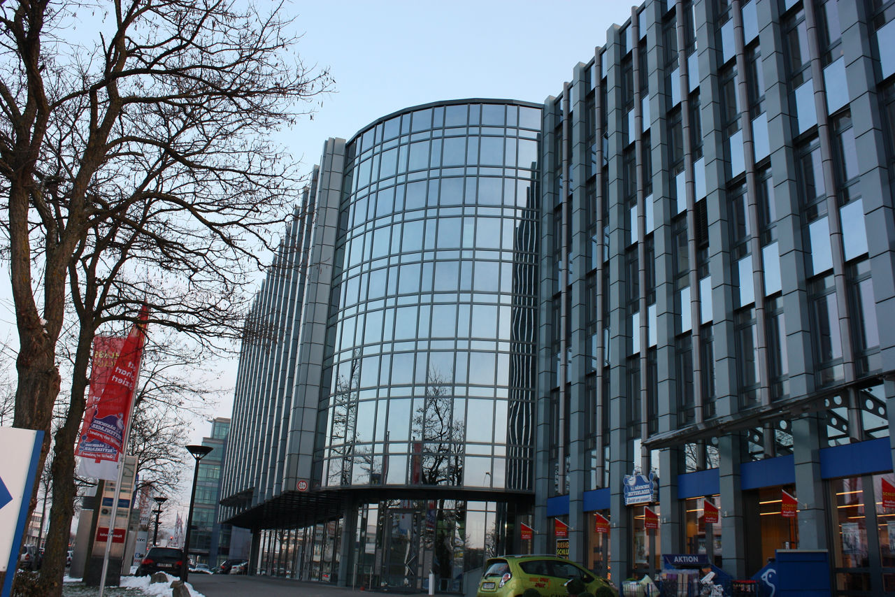 Das Bild zeigt ein modernes Bürogebäude mit einer gebogenen Glasfassade und strukturierten Fensterfronten, vor dem ein Gehweg mit kahlen Bäumen, roten Werbefahnen und einem kleinen grünen Auto verläuft.