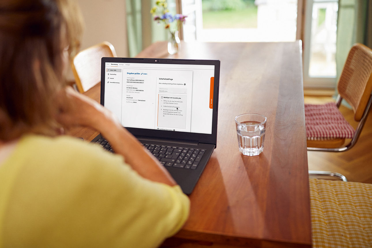 Frau sitzt am Esstisch mit einem aufgeklappten LapTop, auf dem das OnlineBanking zu sehen ist