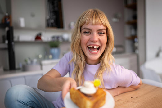 junge Frau mit großer Zahnlücke streckt ein Kuchenstück in die Betrachterrichtung.