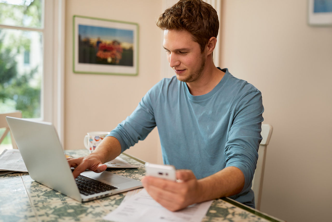 Mann sitzt am Tisch am Laptop und hält sein Handy in der Hand