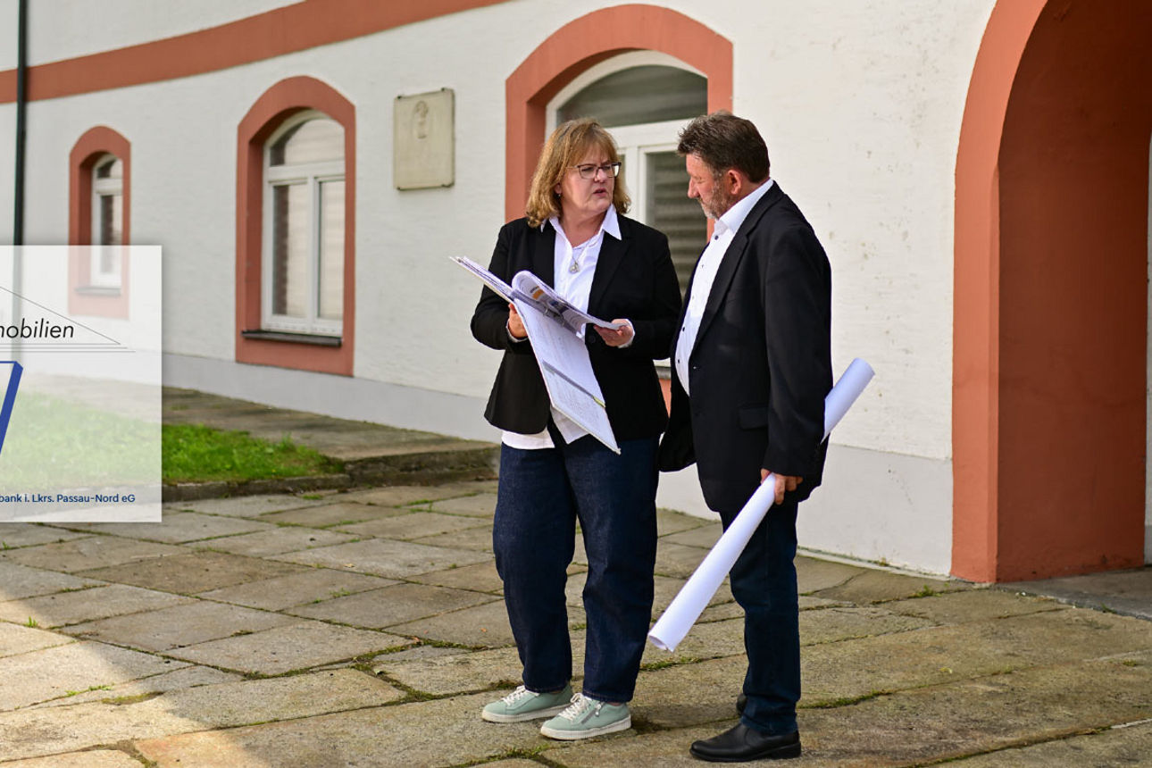 Eine Frau und ein Mann stehen vor einem historischen Gebäude, halten Baupläne in den Händen und unterhalten sich. Links ist d