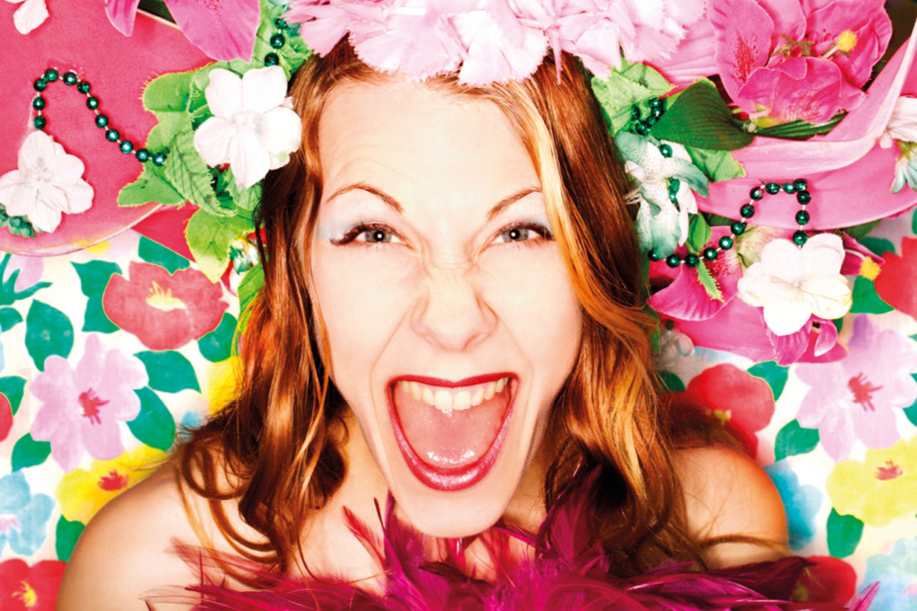 Frau mit Blumenhut lacht ausgelassen vor farbenfrohem Blütenhintergrund.