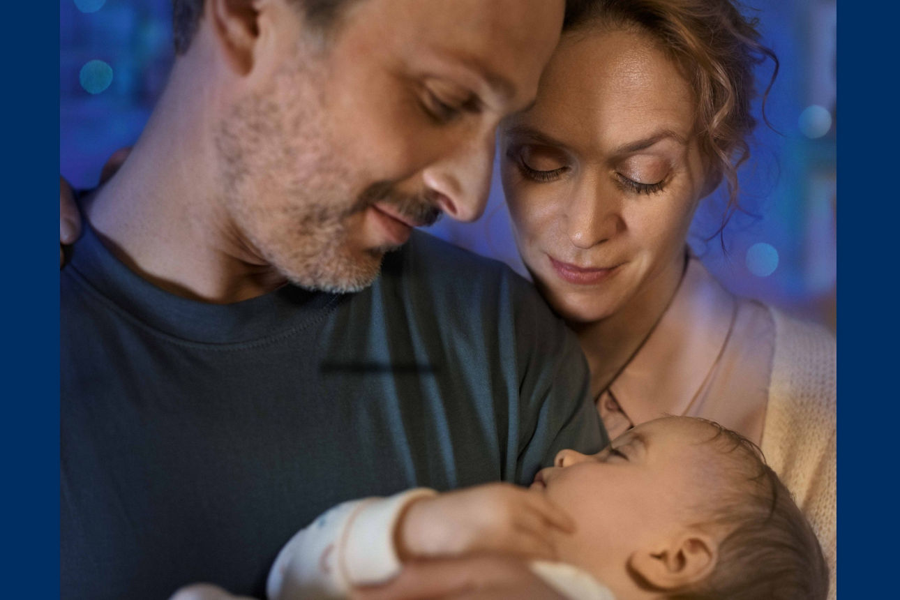 Elternpaar schauen glücklich ihr gemeinsames Baby an, welches der Mann in den Armen hält.