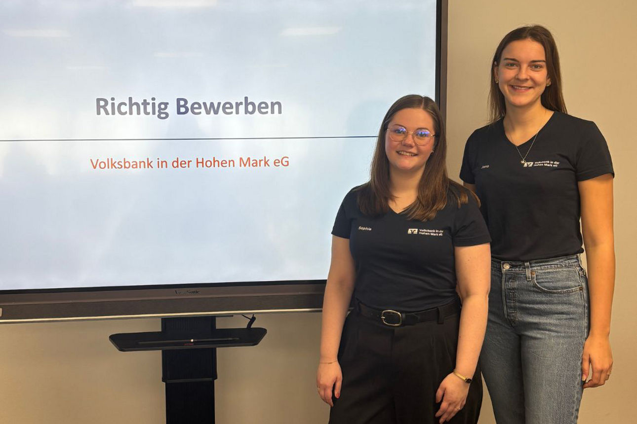 Zwei Mitarbeiter der Volksbank stehen neben einem Präsentationsbildschirm, auf dem ‚Richtig Bewerben‘ sowie ‚Volksbank in der Hohen Mark eG‘ zu lesen ist.