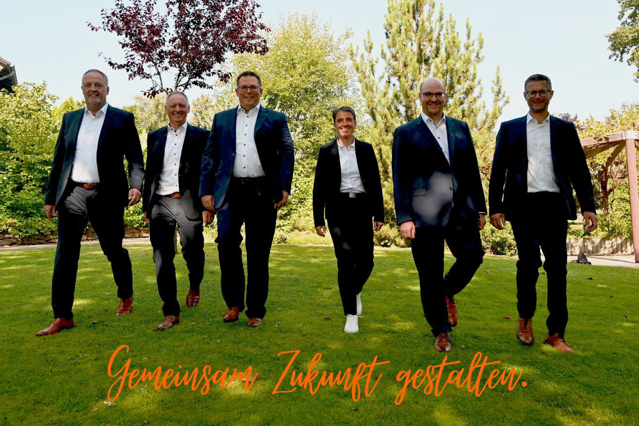 5 Vorstände der Volksbank in der Hohen Mark eG sowie die Generalbevollmächtigte gehen nebeneinander über eine Rasenfläche in einer Parkanlage, alle in dunkler Businesskleidung. Am unteren Bildrand steht der Schriftzug ‚Gemeinsam Zukunft gestalten‘.