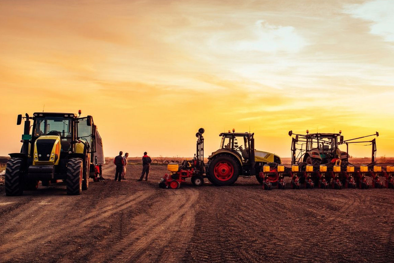 Landwirtschaftliche Maschinen stehen nebeneinander auf einem Feld, darunter Traktoren und ein Sägerät, während mehrere Personen daneben stehen. Im Hintergrund ist ein weiter Acker bei Sonnenuntergang zu sehen.