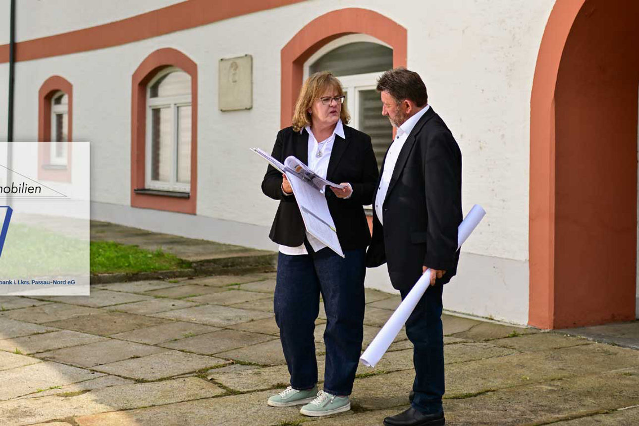Eine Frau und ein Mann stehen vor einem historischen Gebäude, halten Baupläne in den Händen und unterhalten sich. Links ist d