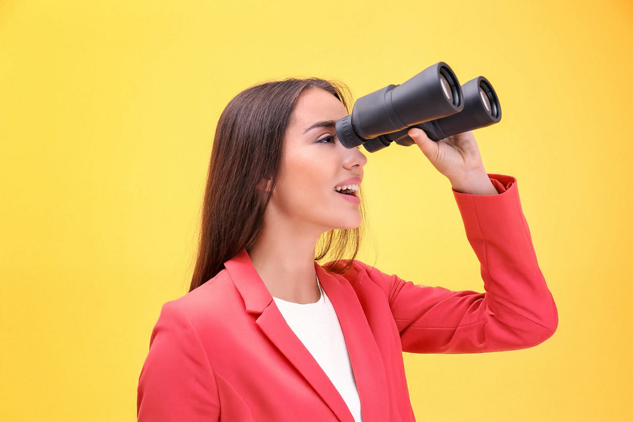 Frau in einem Blazer steht vor einem gelben Hintergrund und schaut durch ein Fernglas