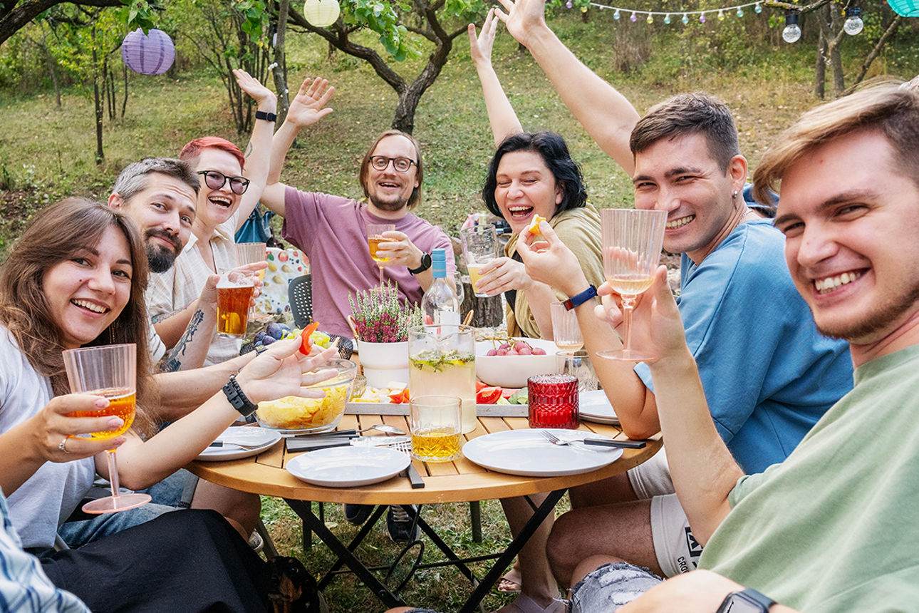 Freundesgruppe grillt im Garten, alle schauen fröhlich zur Kamera und prosten sich zu