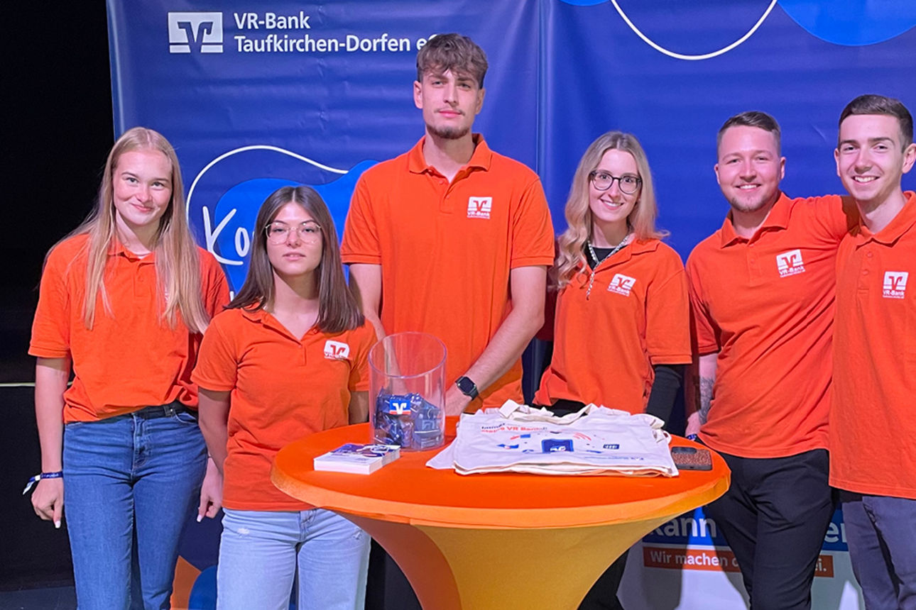 Sechs Kolleginnen und Kollegen stehen in orangen VR-Bank Shirts vor unserer Messewand.