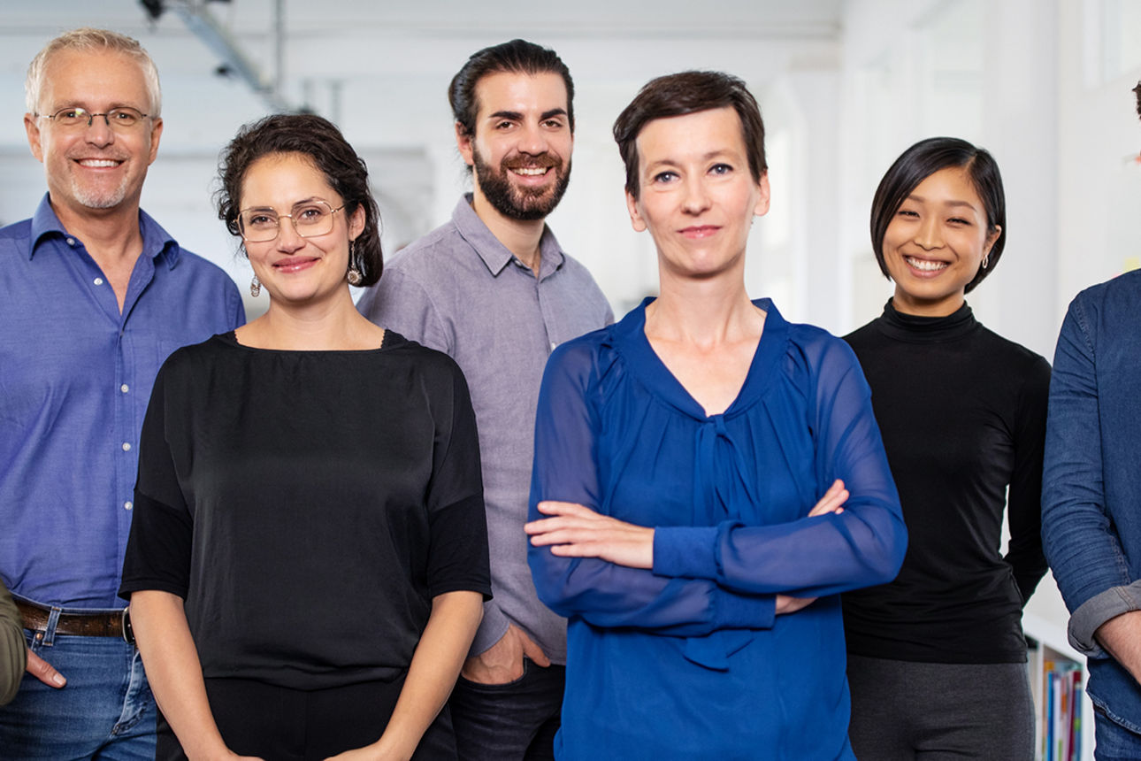 Sieben Personen stehen entschlossen und mit offenem Blick nebeneinander, gekleidet in harmonischen Farben.