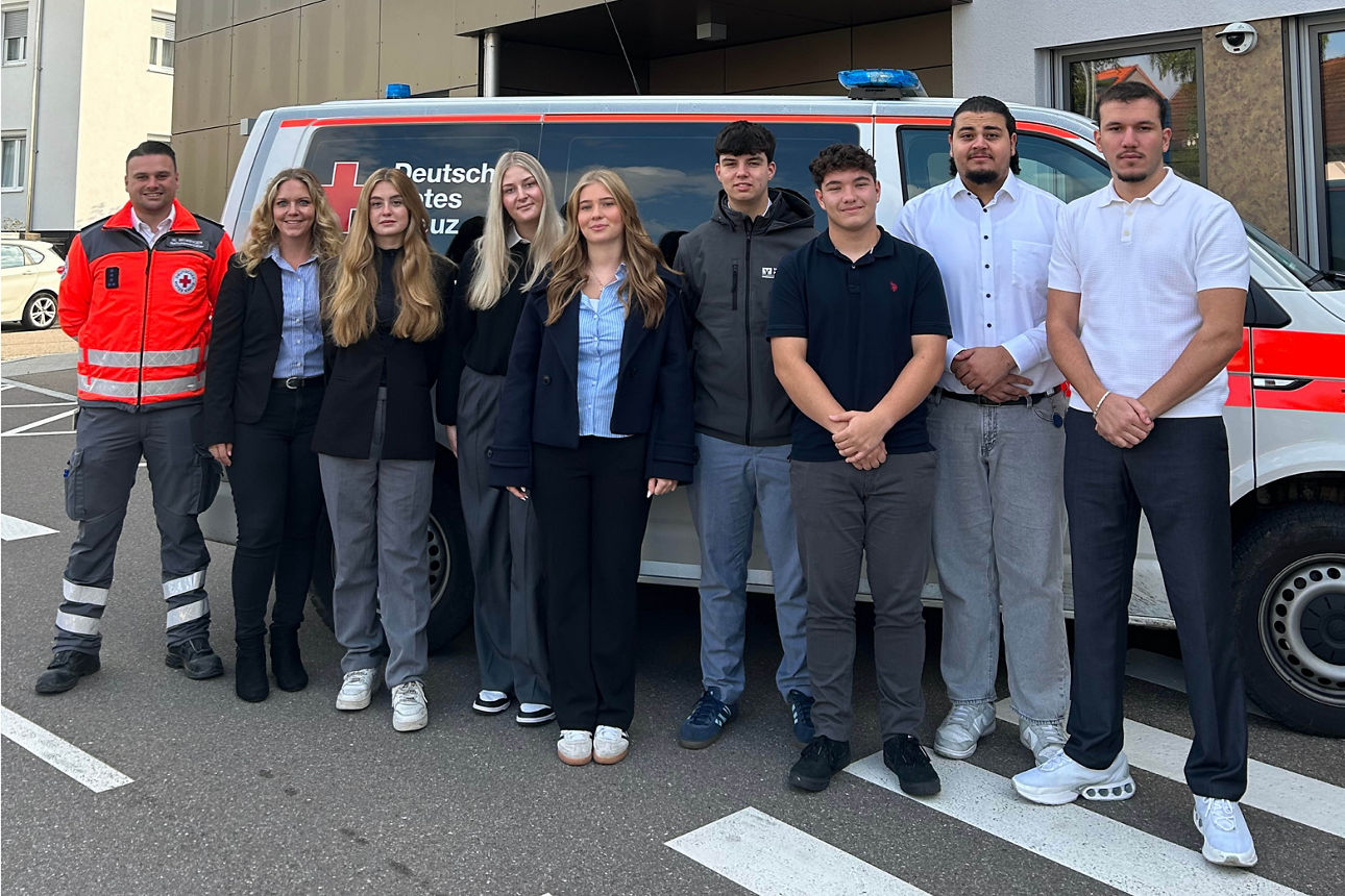 Gruppe von Menschen vor einem Rettungswagen mit der Aufschrift 'Deutsches Rotes Kreuz' vor einem modernen Bürogebäude.