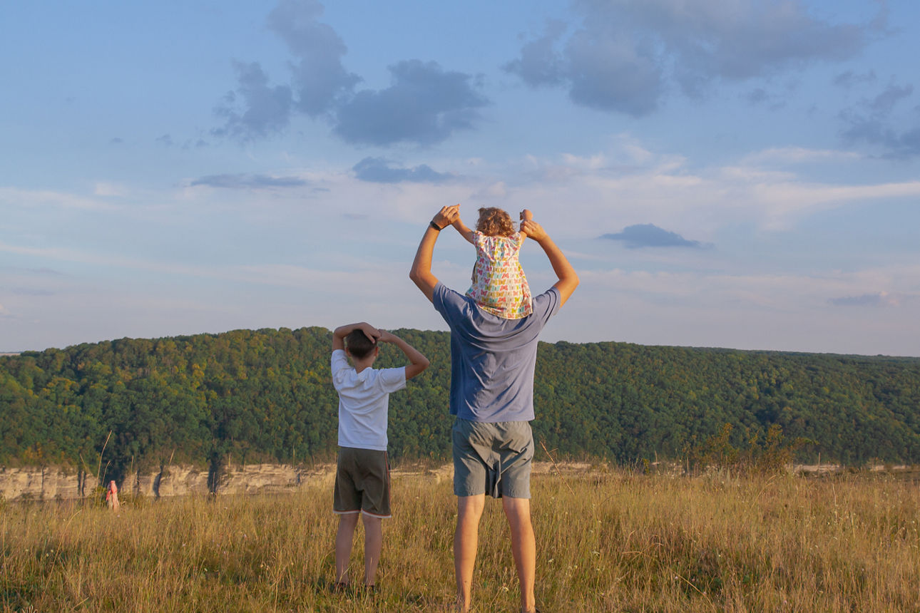 Rückansicht: Vater mit zwei Kindern auf einem Feld, eines der Kinder sitzt auf den Schultern des Vaters
