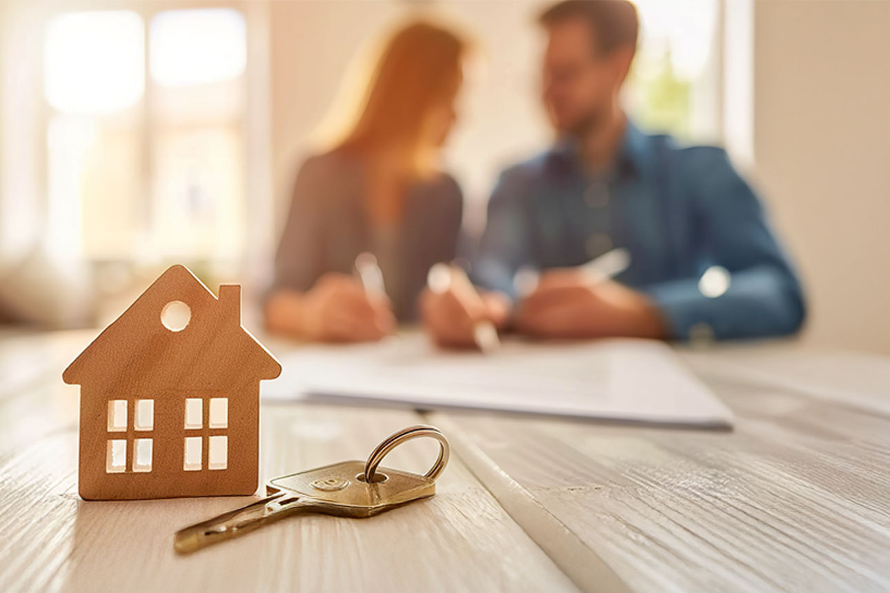Frau und Mann sitzen im Hintergund und im Vordergrund sieht man ein Haus auf den Tisch stehen.
