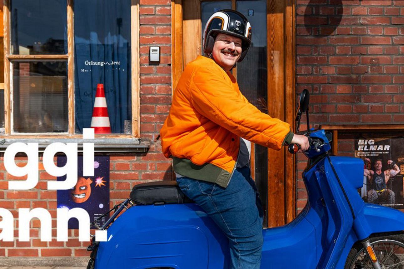 Mann mit Helm und orangefarbener Jacke sitzt auf einem blauen Moped und lächelt in die Kamera