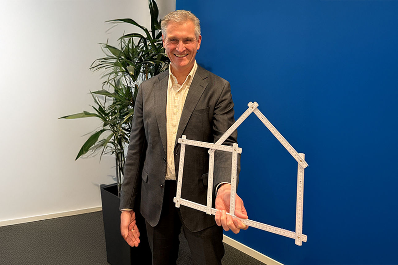 Mann steht vor einer blauen Wand und hält mit einem Zollstock ein Haus in die Höhe