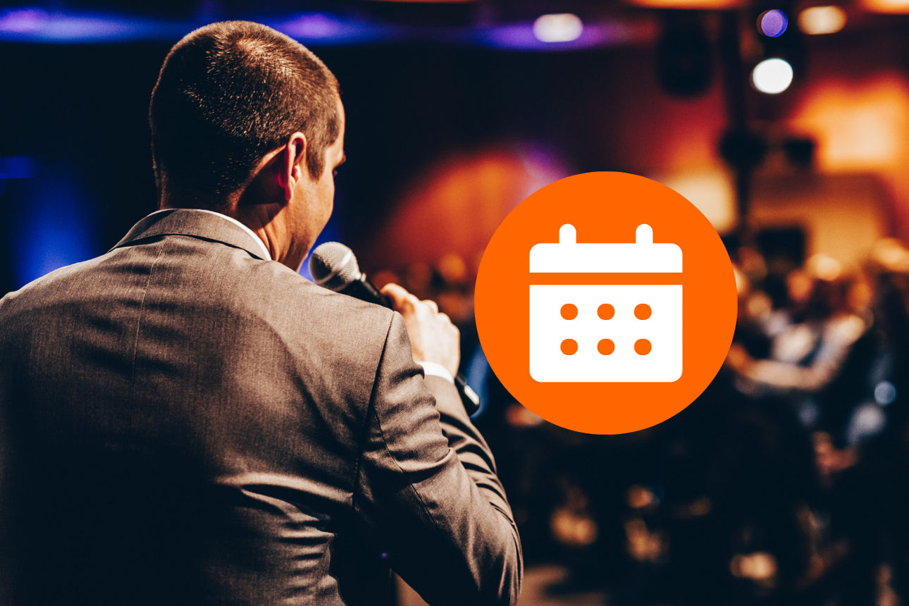 Ein Mann in einem Anzug spricht in ein Mikrofon vor einem Publikum, mit einem Kalender-Symbol in einem orangefarbenen Kreis im Vordergrund..