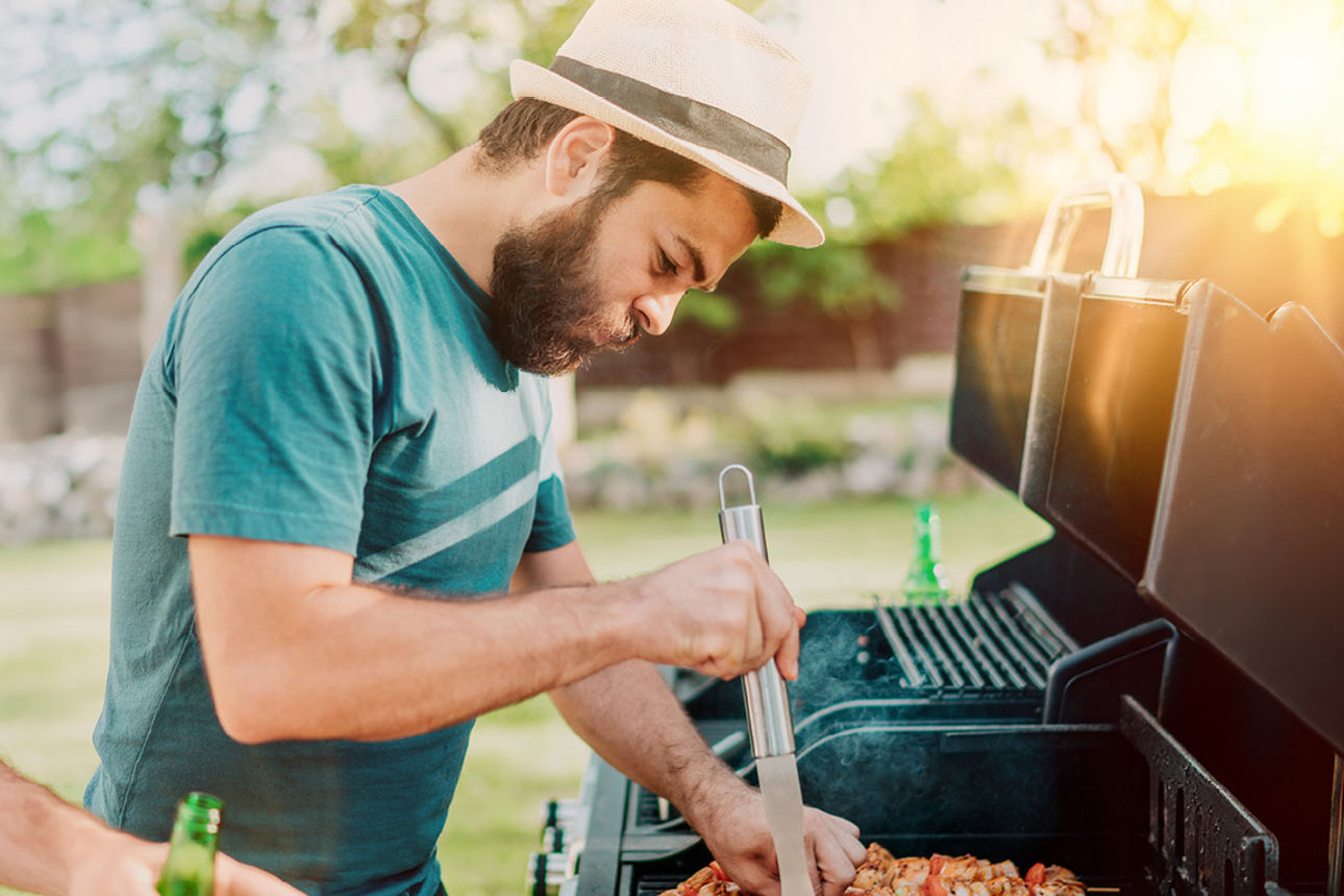 Zwei Männer stehen am Grill im Sonnenuntergang und grillen Schaschlikspieße bei einer Falsche Bier.