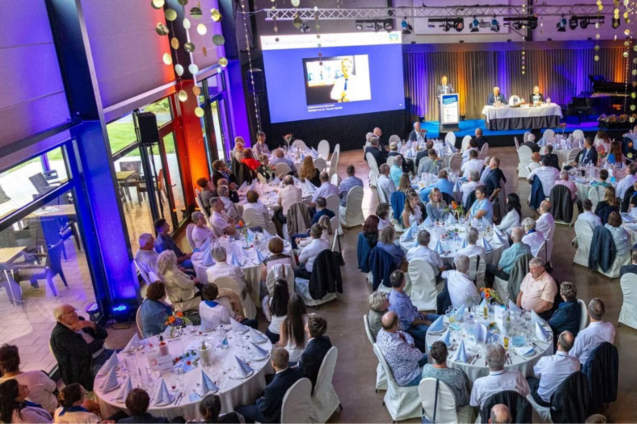 Veranstaltungsszene von oben: rund 100 Personen sitzen an runden Tischen in festlicher Atmosphäre (Vertreterversammlung)