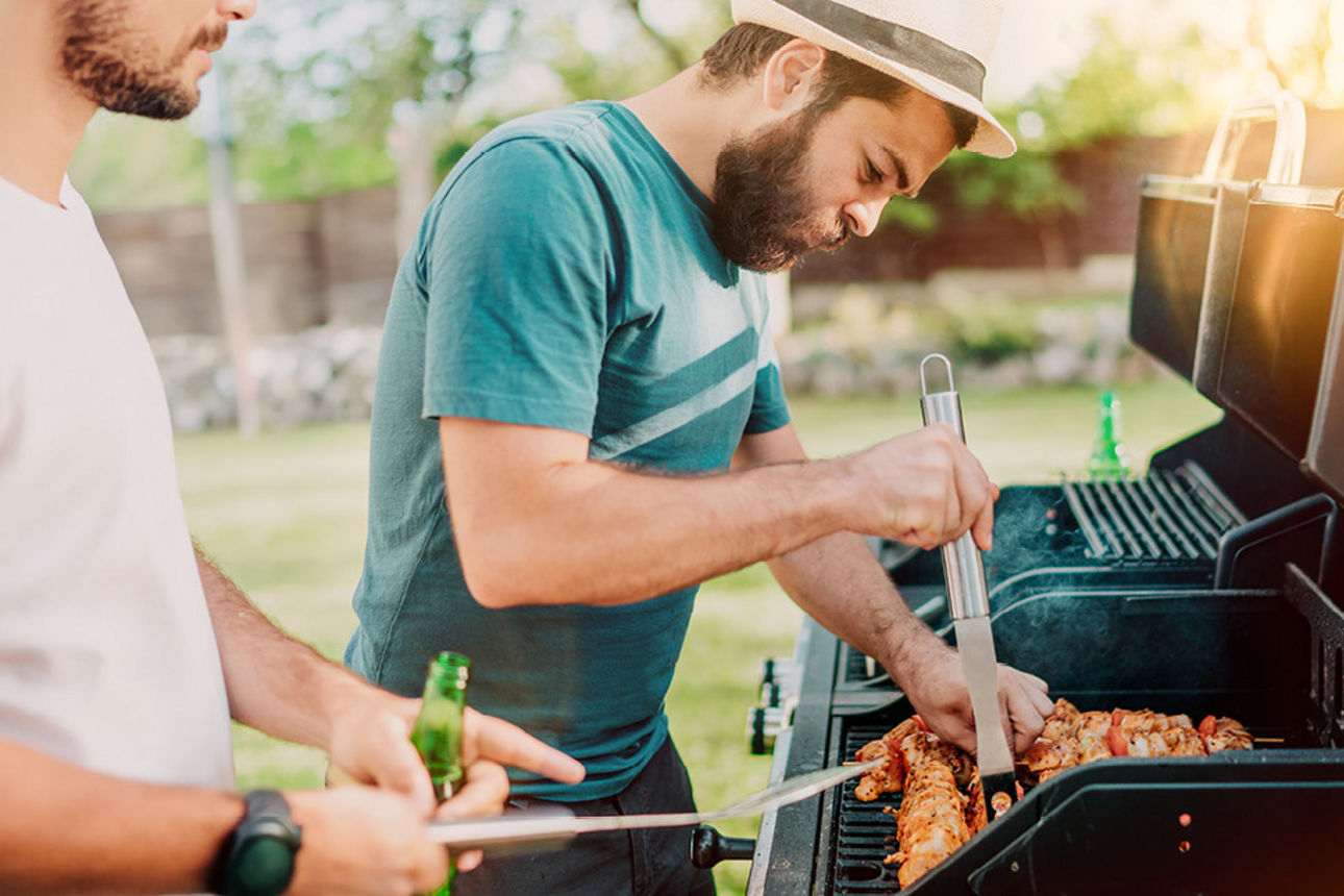 Zwei Männer stehen am Grill im Sonnenuntergang und grillen Schaschlikspieße bei einer Falsche Bier.