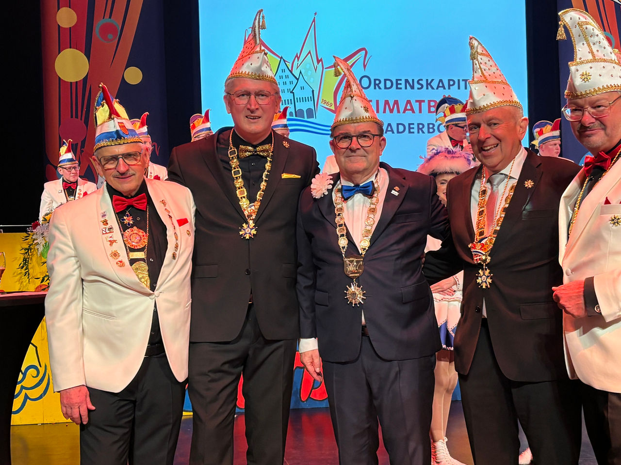 Fünf Männer mit Karnevalsmütze und Karnevalsorden stehen lachend auf einer Bühne.